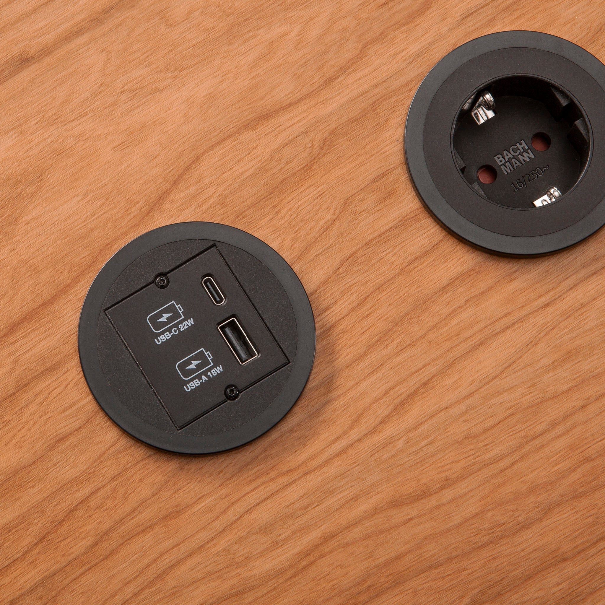 Zwei schwarze, runde Steckdosen sind in eine Holzoberfläche eingebettet; eine verfügt über USB-C- und USB-A-Ladeanschlüsse, die andere ist eine runde Standardsteckdose.