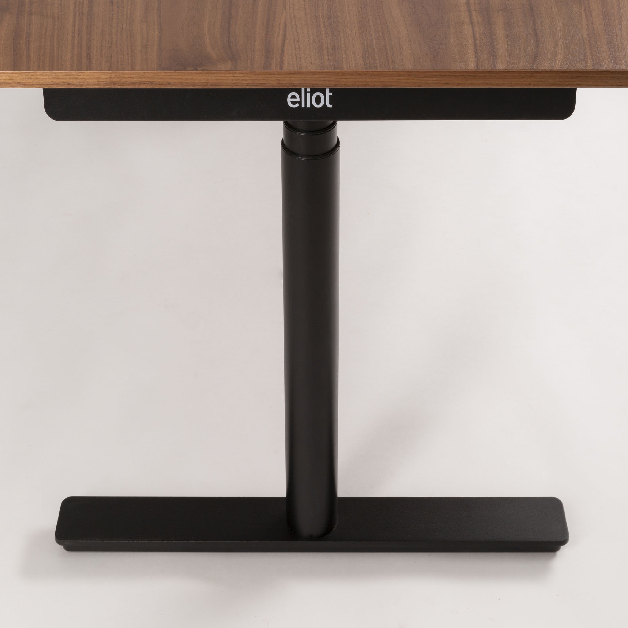 Nahaufnahme eines Holztischs mit schwarzem Metallgestell und -fuß. Auf dem Metallgestell unter der Tischplatte ist das Wort Eliot aufgedruckt. Der Hintergrund ist einfarbig und hell.
