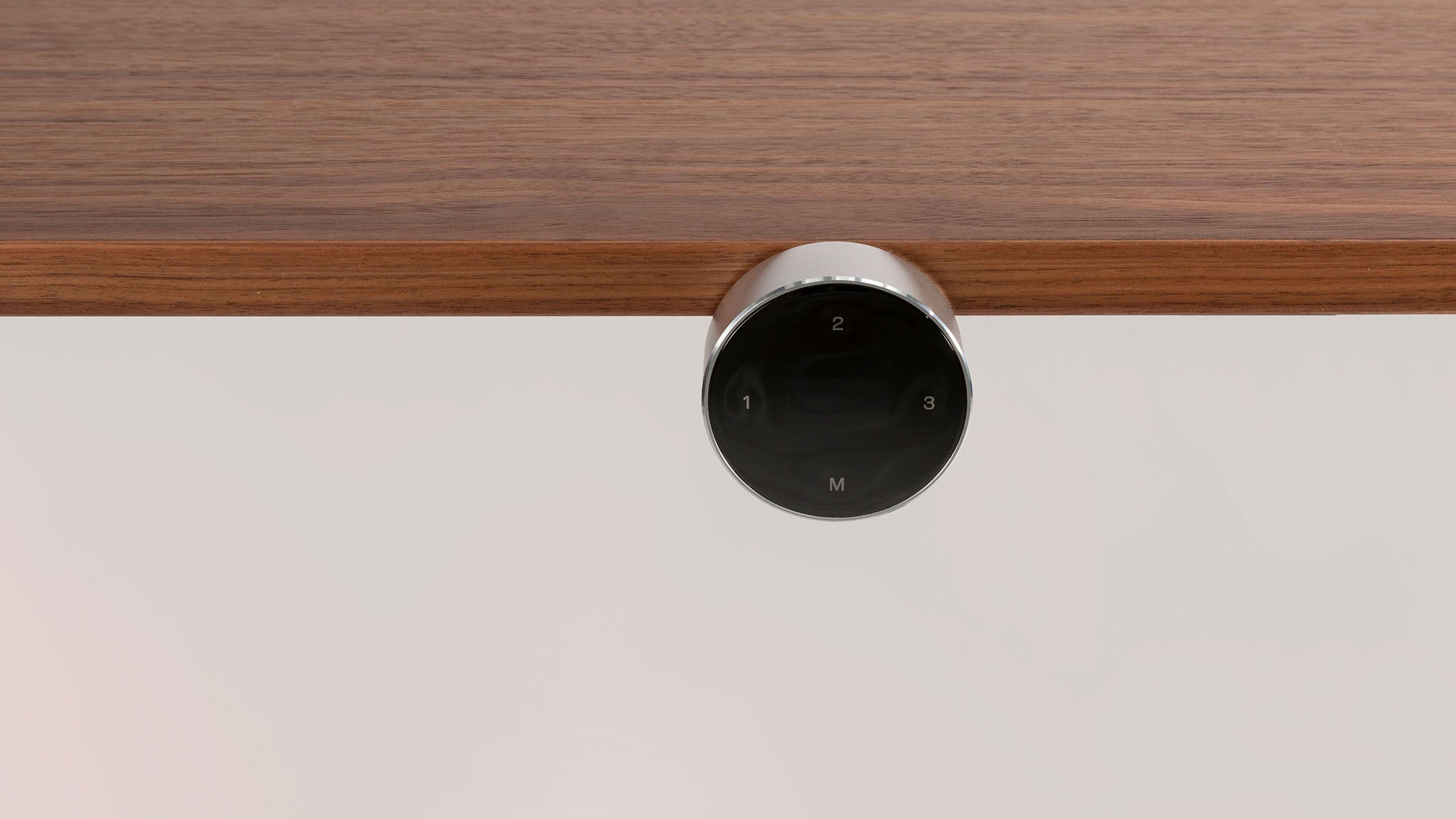 A round, black control knob with numbers and a letter M is mounted under a wooden surface, likely for adjusting settings on a piece of furniture.