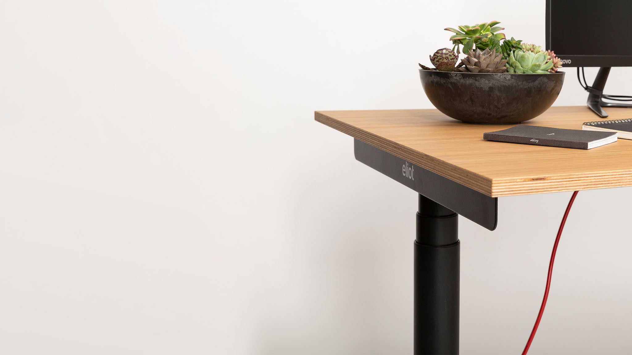A modern wooden desk with black legs holds a black bowl of succulents, a closed notebook, and part of a computer monitor against a plain white wall. A red cable hangs below the desk.