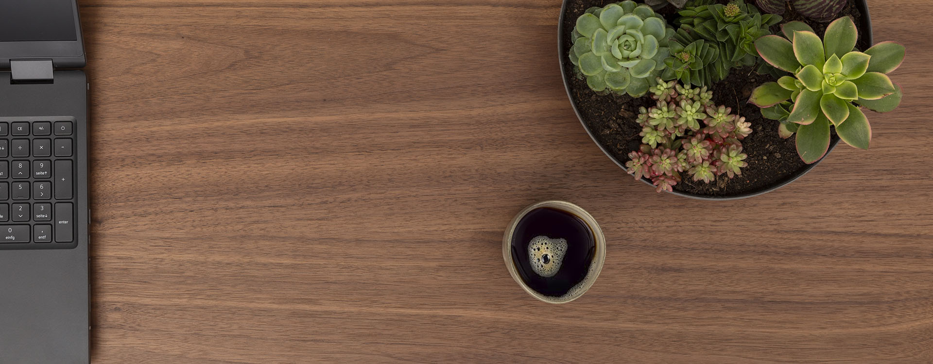 A wooden desk with a laptop keyboard on the left, a small bowl of black coffee in the center, and a pot of various succulents on the upper right. The central area of the desk is clear.