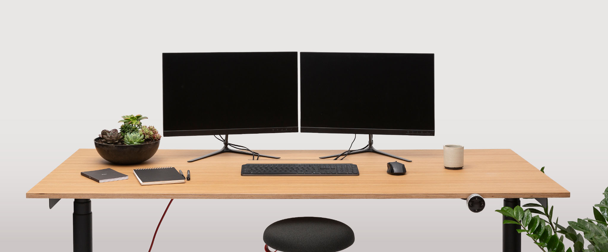 A tidy wooden desk with two black monitors, keyboard, mouse, notebooks, pen, succulents bowl, small speaker, and floor plant on the right. A round black stool sits in front—the PRO is the perfect place to focus and pursue your mission.
