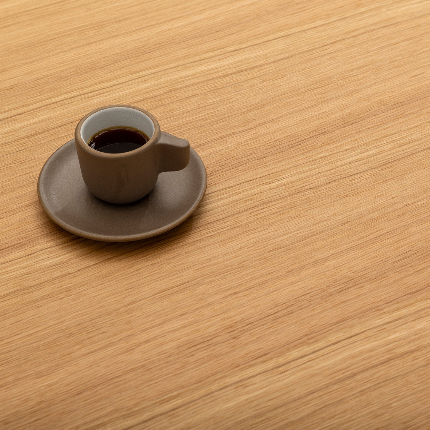 A small brown cup filled with coffee sits on a matching saucer, placed on a light wooden surface with a visible grain pattern. The cup is positioned in the upper left area of the image, leaving ample empty space.