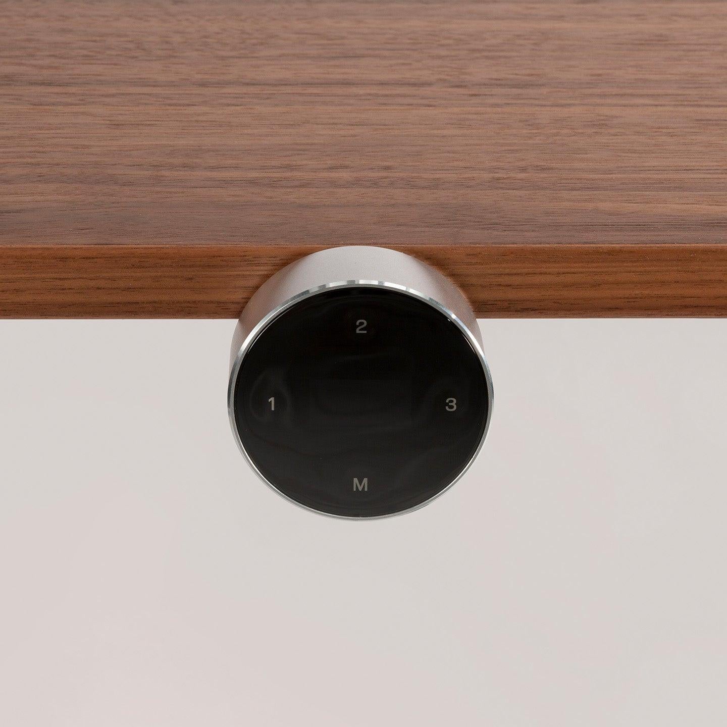 Eine Nahaufnahme des elektronischen EXPAND-Bedienfelds von CUBE Products & Furniture mit vier Touch-Tasten (1, 2, 3, M) in Schwarz und Silber, die unter einer Holzoberfläche angebracht sind - ideal für jede hochwertige oder moderne Arbeitsplatzeinrichtung.
