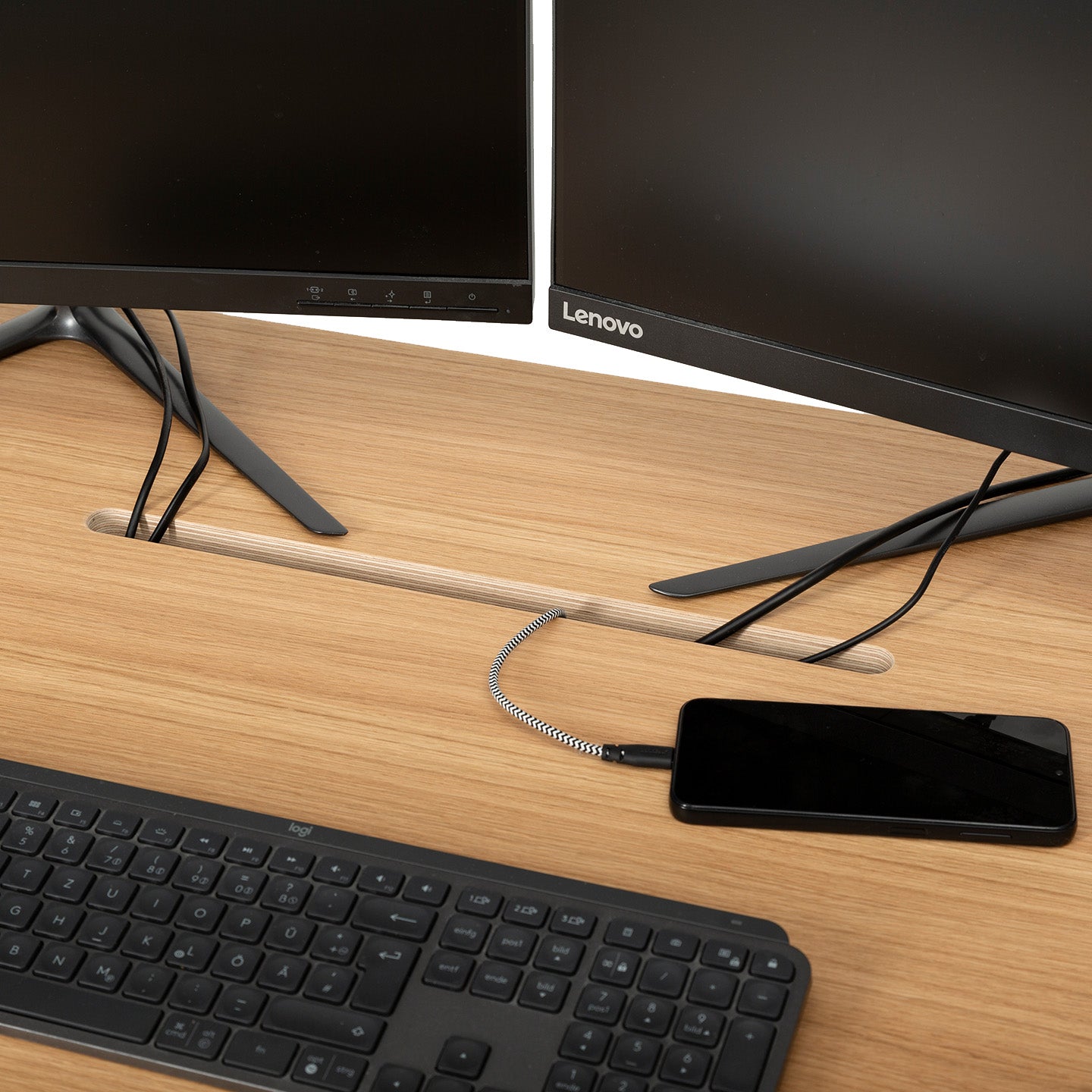 A wooden desk made from MDF Platten includes a black keyboard, a coiled-cable smartphone, two Lenovo monitors, and neatly managed cables passing through a Kabeldurchlass - Oblong gefräst slot.