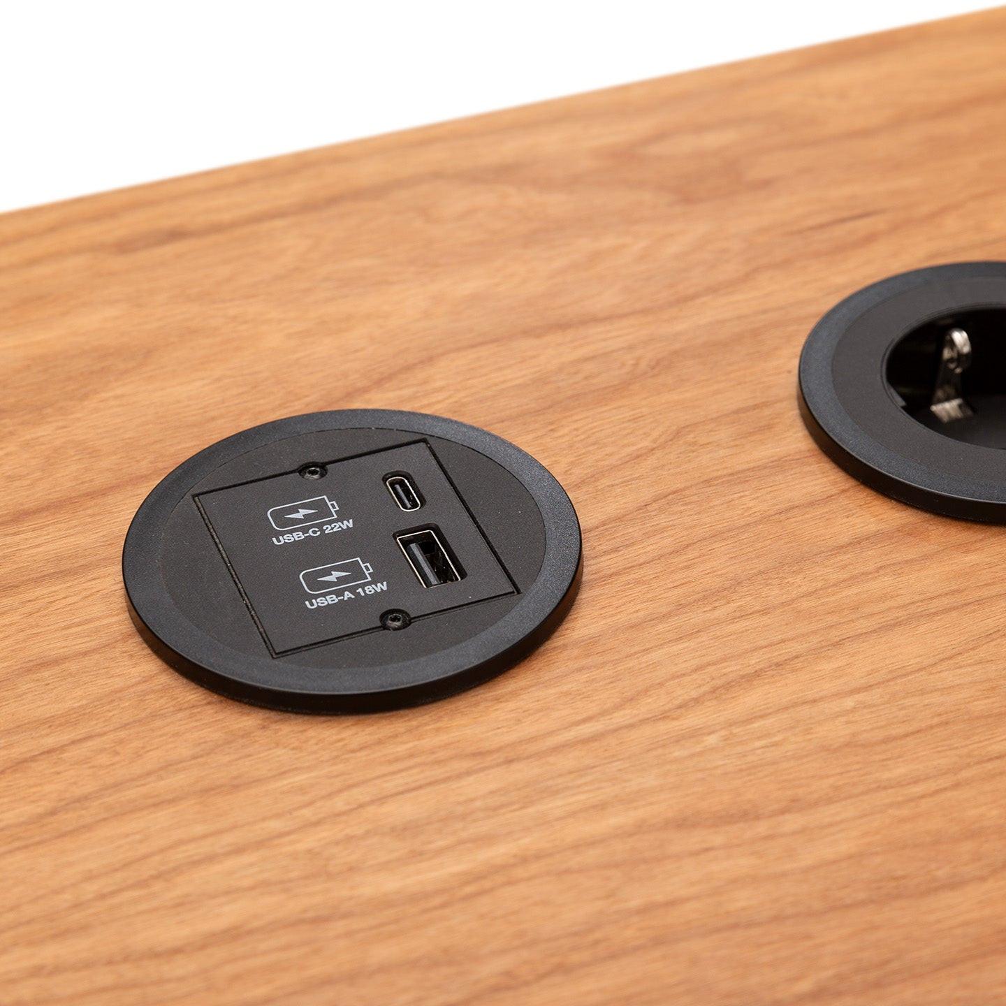 Nahaufnahme der hölzernen Arbeitsfläche MAX von CUBE Products & Furniture mit integrierter Steckdose und beschrifteten USB-C- und USB-A-Anschlüssen, umgeben von einer schwarzen, runden Umrandung - perfekt für besonders große Setups mit mehreren Monitoren.
