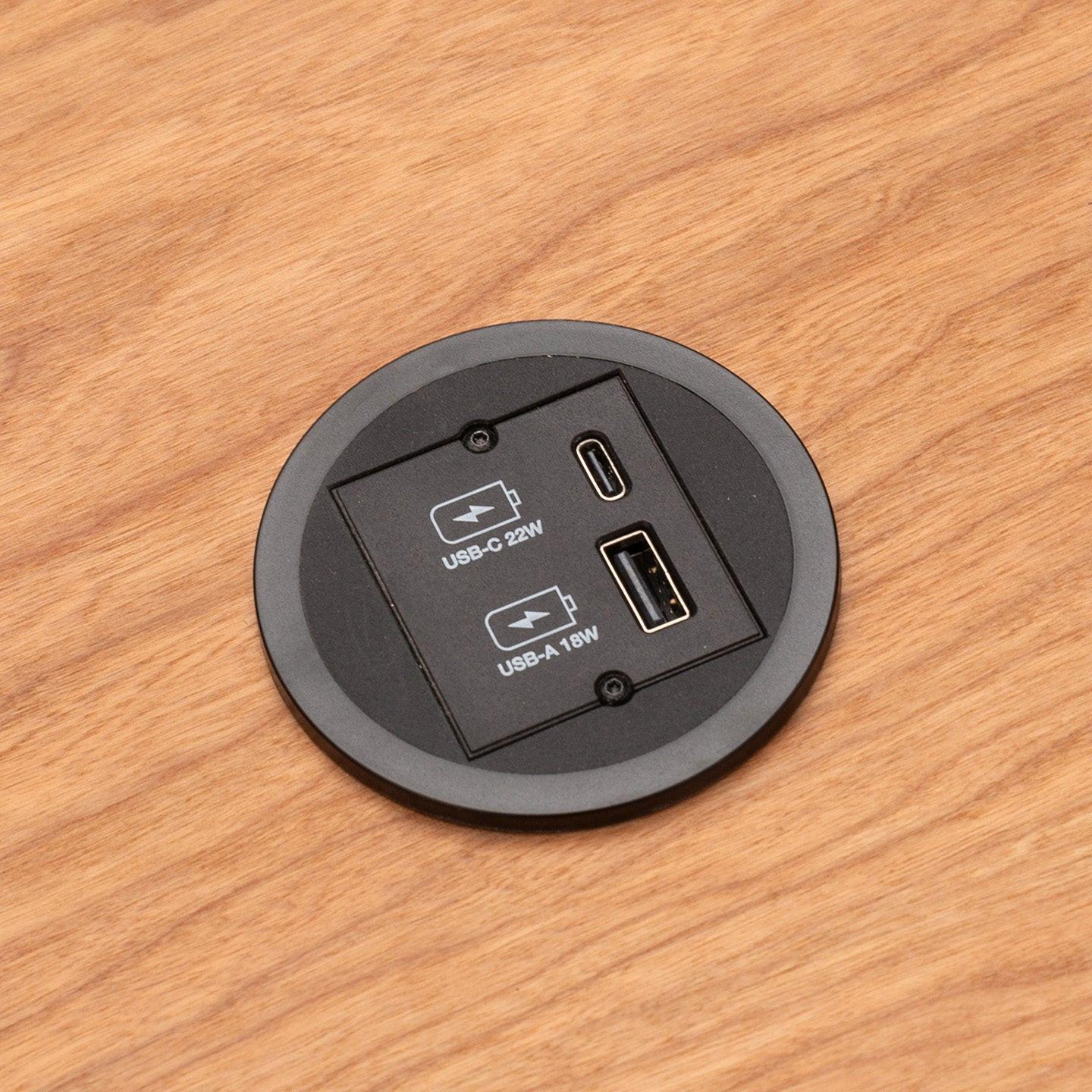 Eine Nahaufnahme eines CUBE Products & Furniture Kombicharger USB A/C mit eingebautem rundem Modul auf einem Holztisch, mit einem 22W USB-C Power Delivery-Anschluss und einem 18W USB-A-Anschluss.