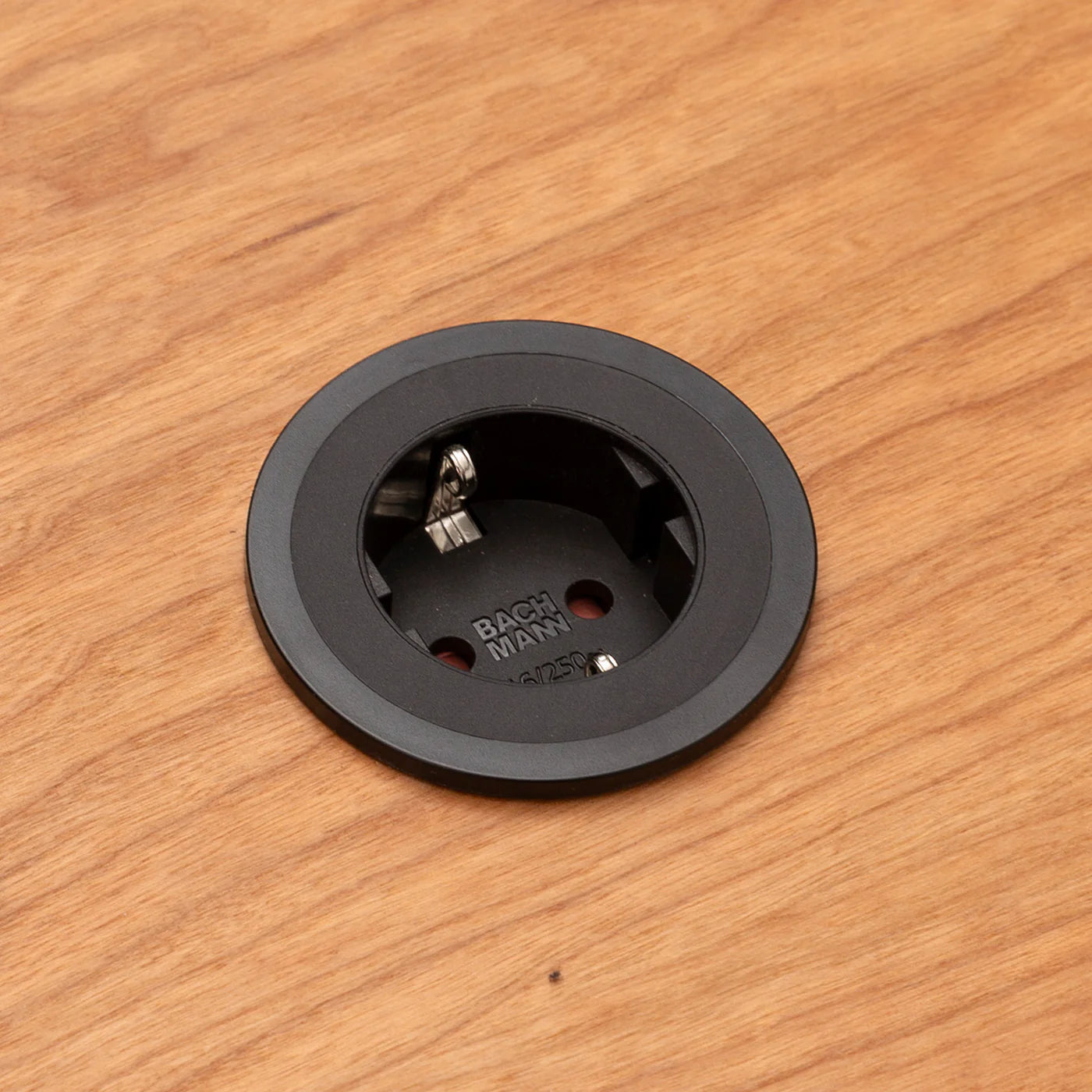 Eine runde, schwarze FOCUS-Steckdose von CUBE Products & Furniture ist von oben gesehen in einen Holztisch eingelassen. Das versenkte Design mit zwei Befestigungsschrauben eignet sich perfekt für kompakte Arbeitsbereiche oder ein modernes Home Office.