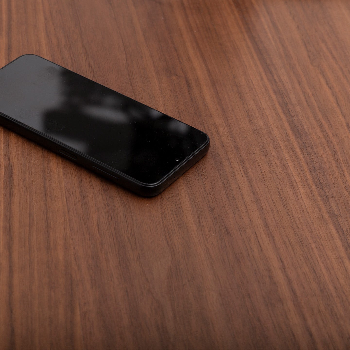 A black smartphone rests face up on the left side of a wooden surface, its dark screen catching the light atop a Tischplatte - Furnier Walnuss, while the elegant walnut veneer table remains empty.