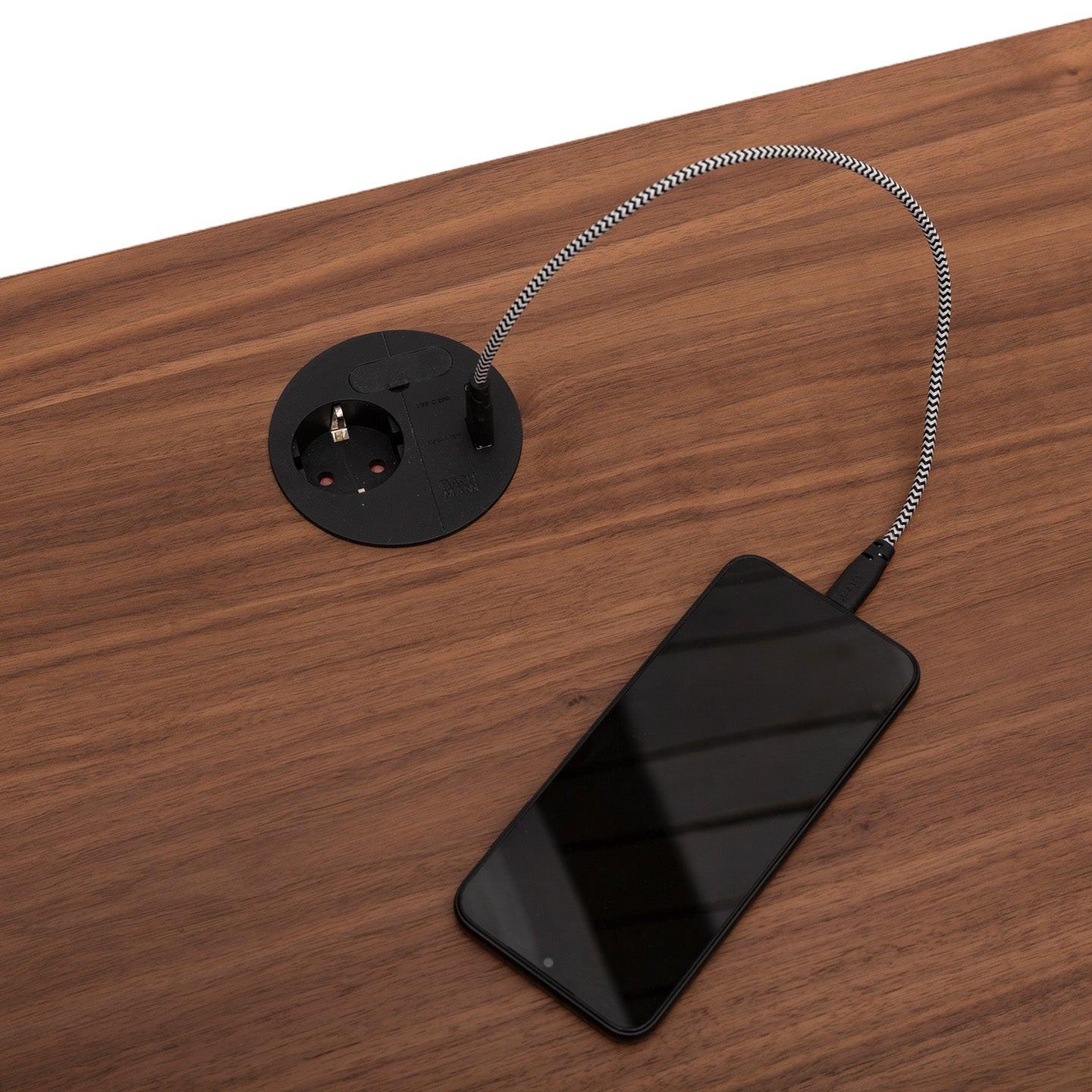 Ein Smartphone wird an ein schwarz-weißes, geflochtenes Ladekabel angeschlossen, das in eine runde MAX-Steckdose von CUBE Products & Furniture eingesteckt wird, die in eine hölzerne Tischplatte eingelassen ist - perfekt für XL-Setups oder große Arbeitsbereiche mit mehreren Monitoren.
