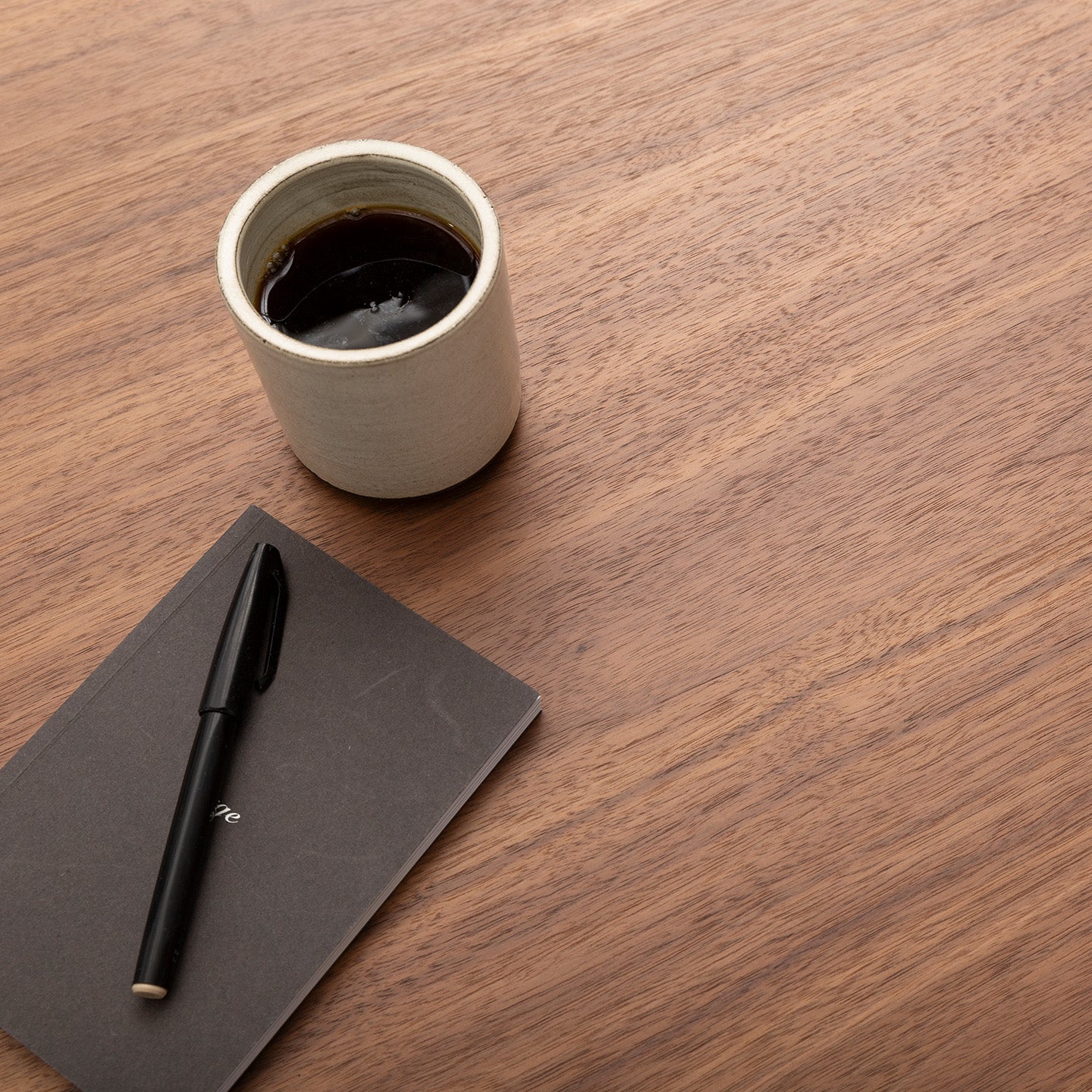 A ceramic cup of black coffee rests on a Tischplatte - Furnier Walnuss, beside a closed black notebook with a pen on top.