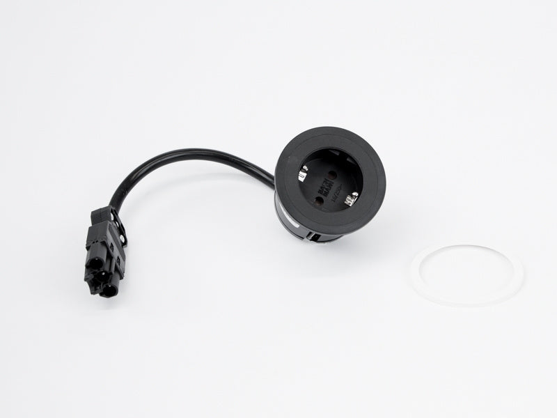 A black round Einbausteckdose Pix with a GST 18 Zuleitung cable and connector rests on a white surface beside a small white circular ring.