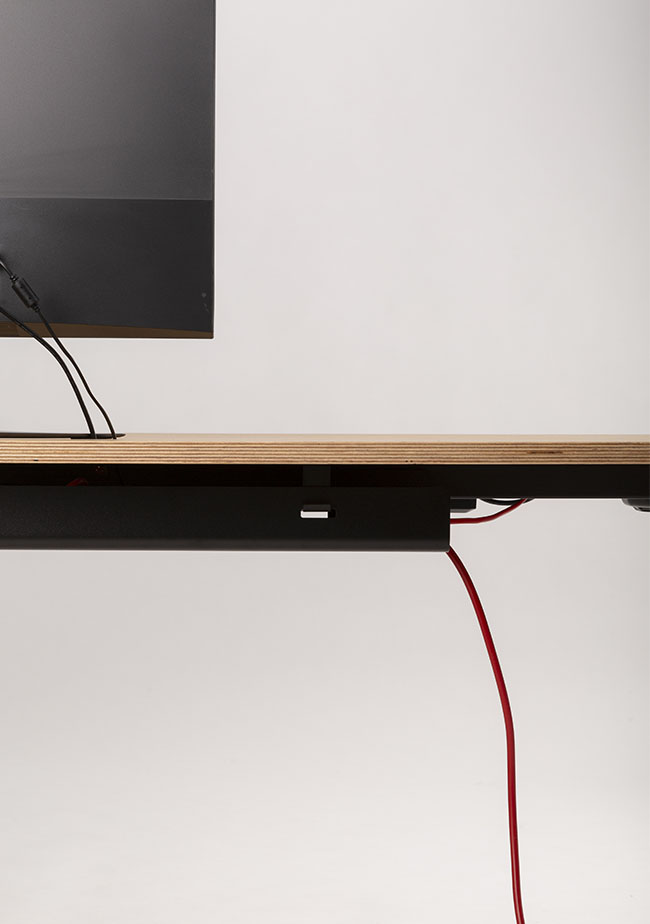 Minimalist side view of a wooden desk with a black monitor partially visible. Red and black cables, organized with the Kabelwanne - 1 Cable PRO, extend from the desk against a light gray background.
