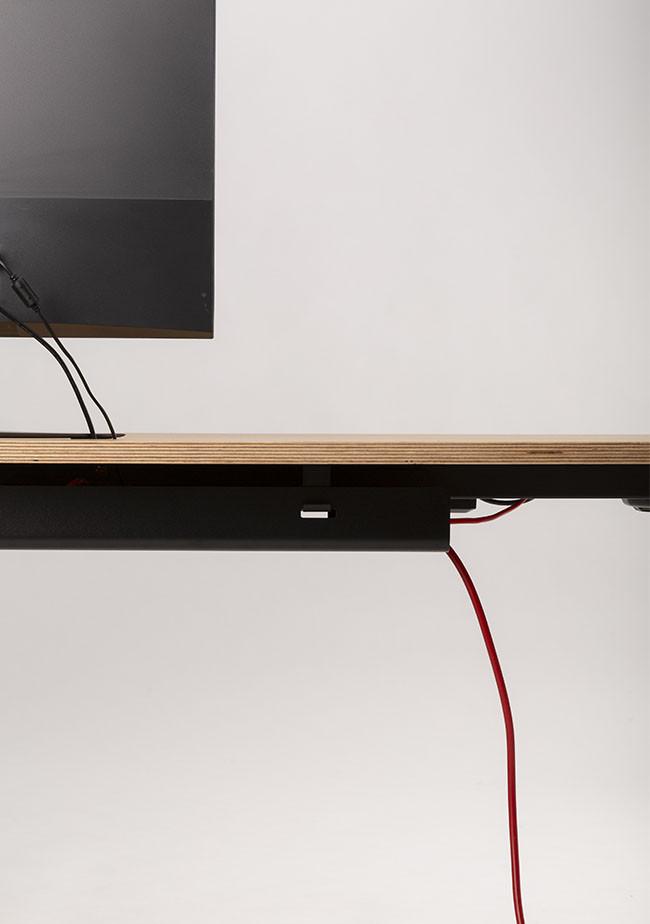 Der MAX-Schreibtisch von CUBE Products & Furniture hat eine Holzoberfläche mit schwarzer Kante, die einen Monitor und sichtbare Kabel (auch rote) trägt und ein XL-Setup und einen organisierten Arbeitsbereich vor einem schlichten Hintergrund hervorhebt.