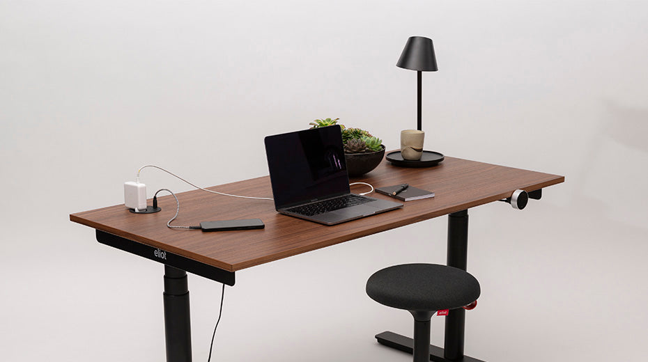 A modern wooden desk with a black laptop, smartphone, charging devices, a lamp, a plant, a mug, and a black swivel stool against a plain white background.