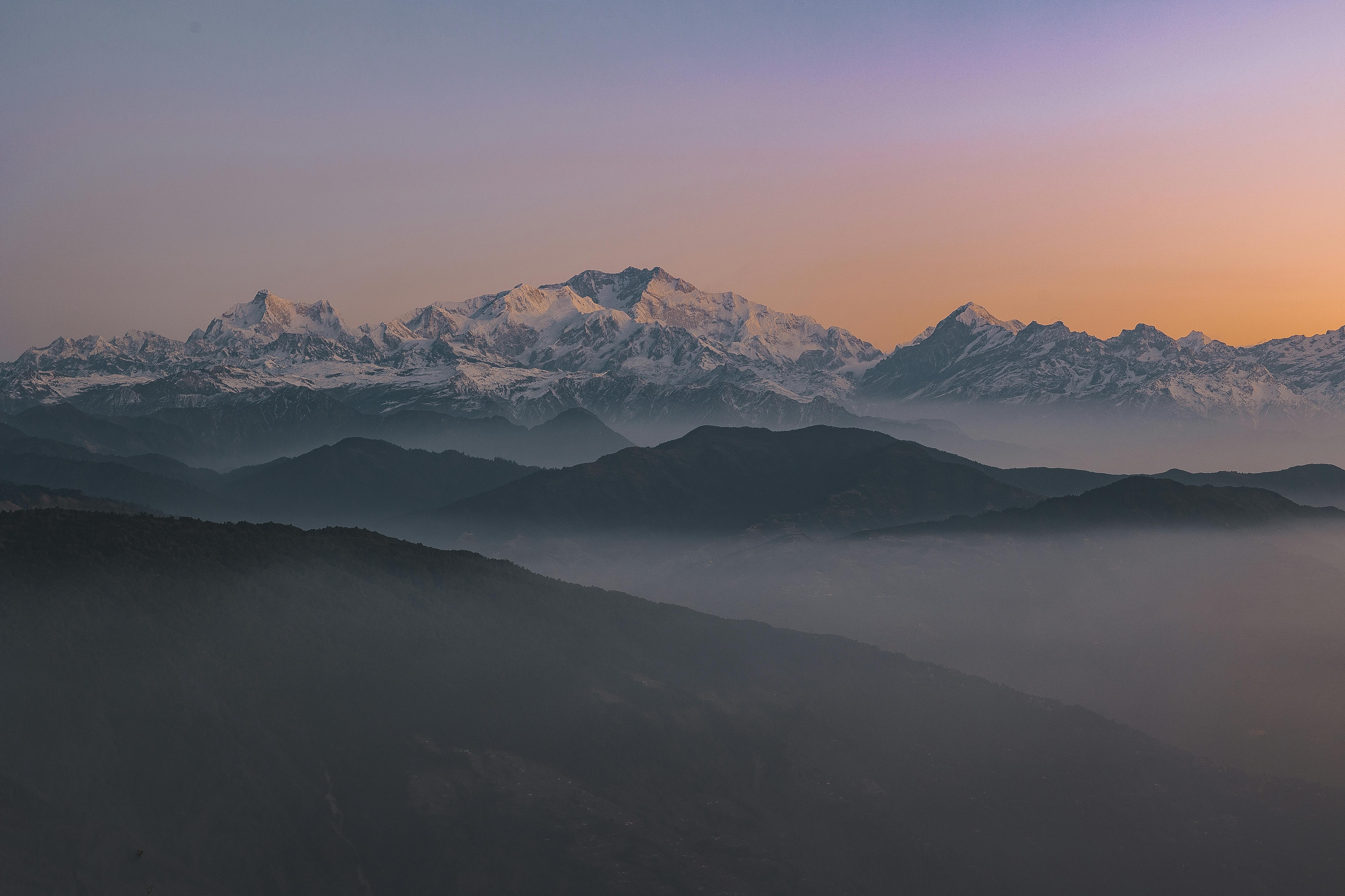 Snow-capped mountains under a clear sky at sunrise or sunset, with layers of dark hills and mist in the foreground, creating a serene and tranquil landscape.