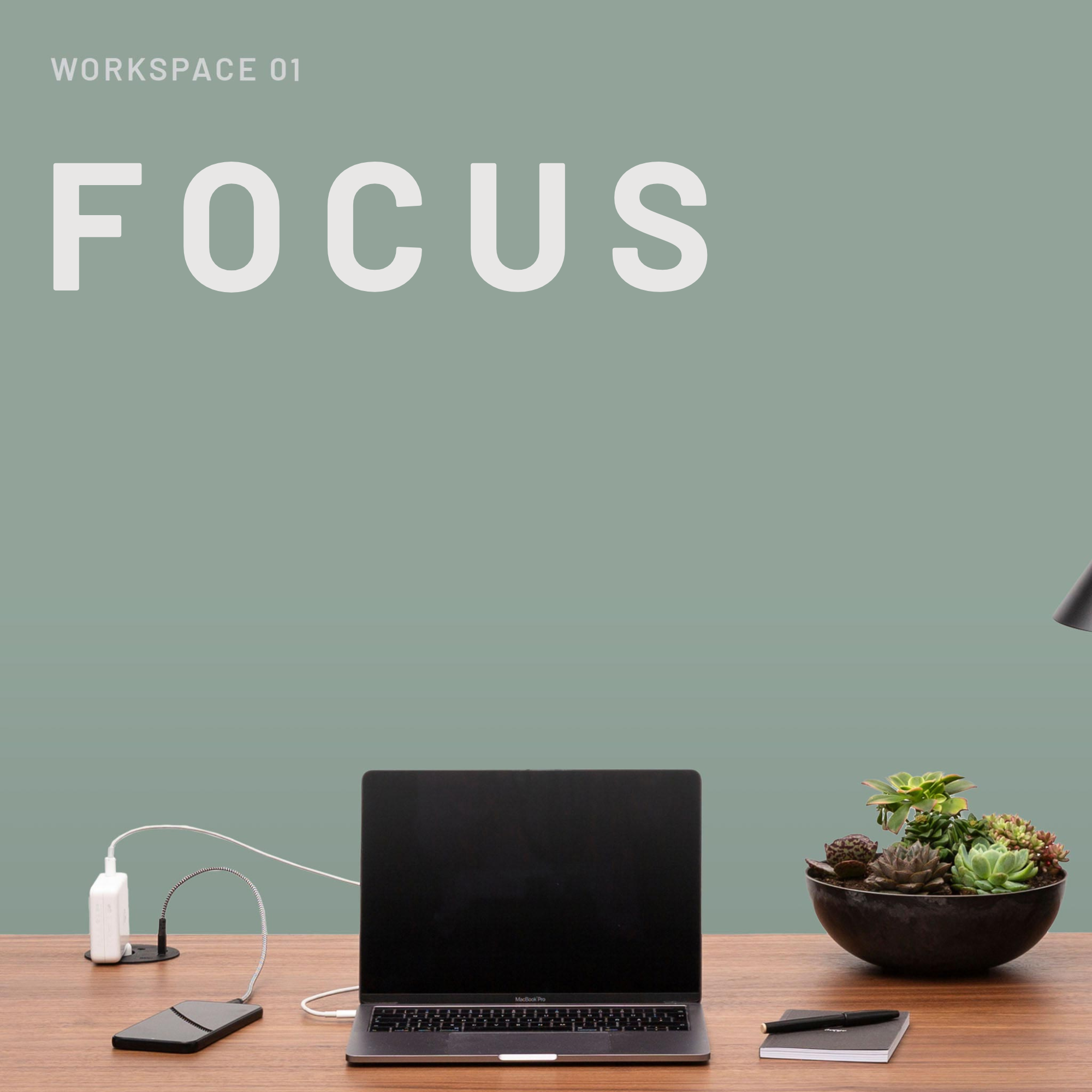 Arbeitsbereich 01: FOCUS von CUBE Products & Furniture - ein minimalistischer Home-Office-Schreibtisch mit MacBook Pro, Smartphone, Ladekabel, Sukkulentenschale, Notizbuch und Stift vor einer salbeigrünen Wand.