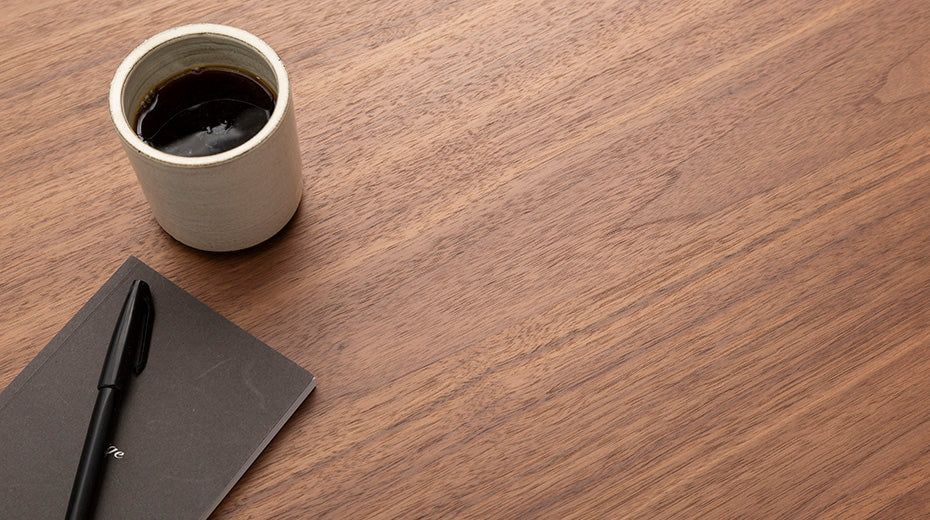 A ceramic cup of black coffee sits on a wooden table beside a dark notebook and a black pen. The table surface is clean and uncluttered, with ample empty space visible.