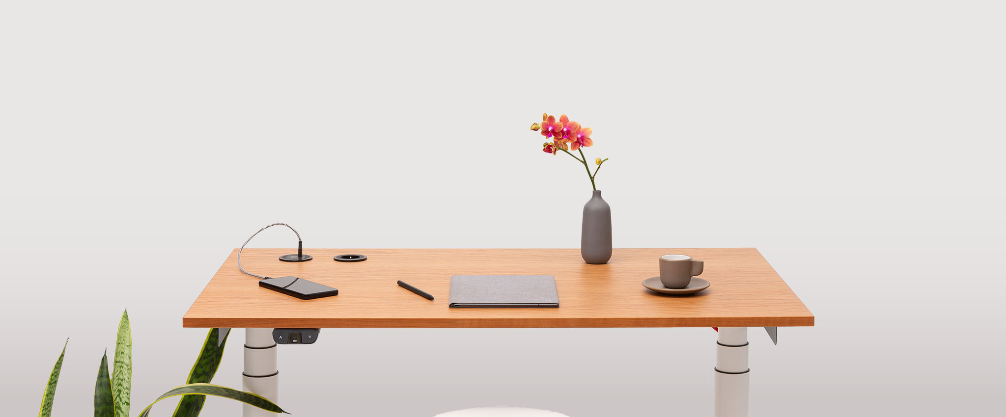 The TINY desk features a smartphone, pen, notebook, gray vase with pink flowers, and a cup and saucer—your perfect Begleiter im Alltag. A green plant is partially visible in the lower left against a simple background.
