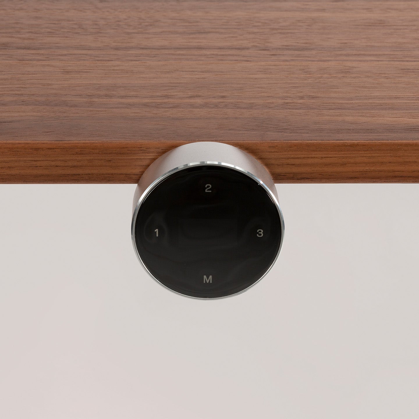 The Smartcontroller - Eliot, a round black controller with a silver rim, is mounted under a wooden surface. Its panel shows 1, 2, 3, and M—ideal for your höhenverstellbarer Schreibtisch setup.