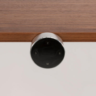Eine Nahaufnahme des elektronischen EXPAND-Bedienfelds von CUBE Products & Furniture mit vier Touch-Tasten (1, 2, 3, M) in Schwarz und Silber, die unter einer Holzoberfläche angebracht sind - ideal für jede hochwertige oder moderne Arbeitsplatzeinrichtung.