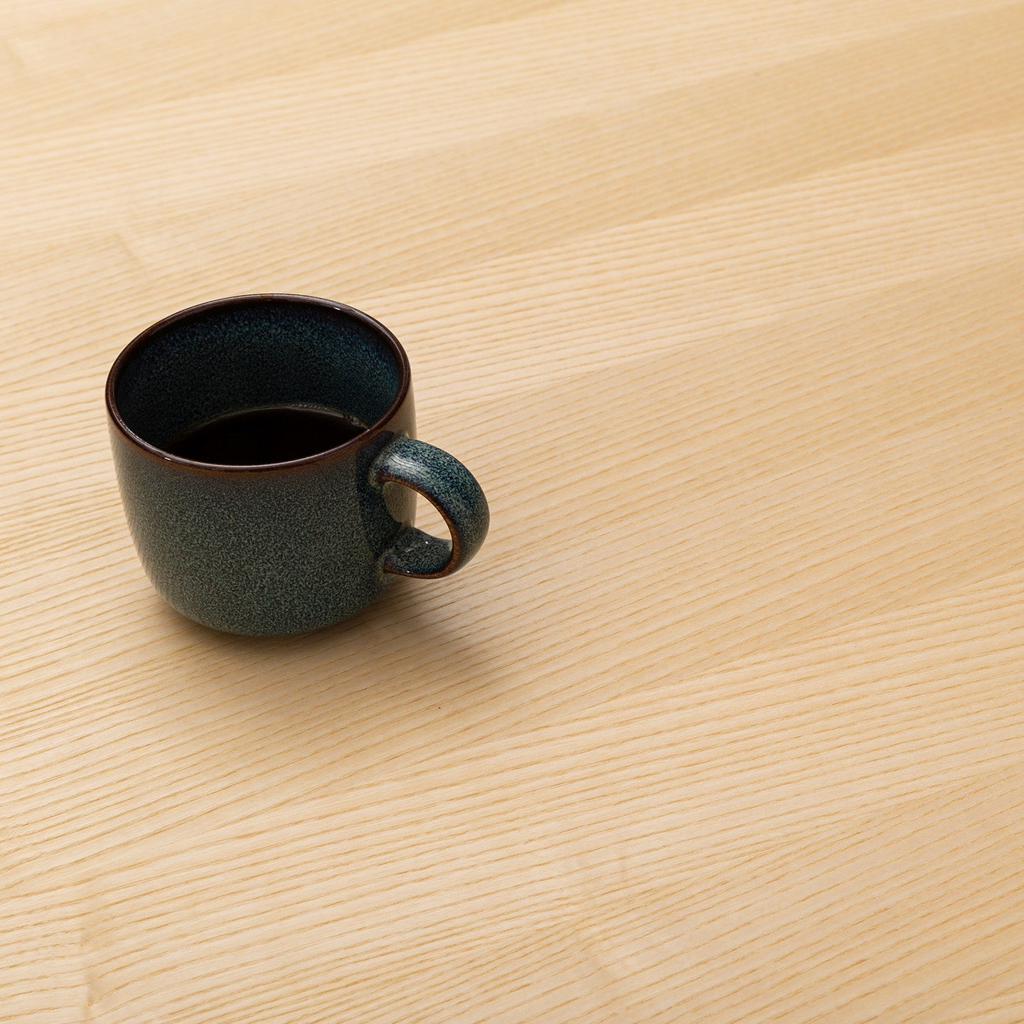 A FLEX dark ceramic coffee cup filled with black coffee sits on a light wooden table in a cozy home office, positioned toward the left side of the otherwise empty surface.