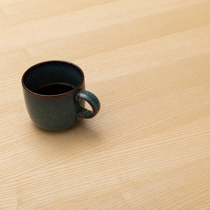 A FLEX dark ceramic coffee cup filled with black coffee sits on a light wooden table in a cozy home office, positioned toward the left side of the otherwise empty surface.