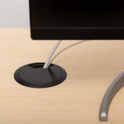 A close-up of a computer monitor on a light wood desk, with two white cables passing through a Kabeldurchlass - Kunststoff in the desktop for organization.