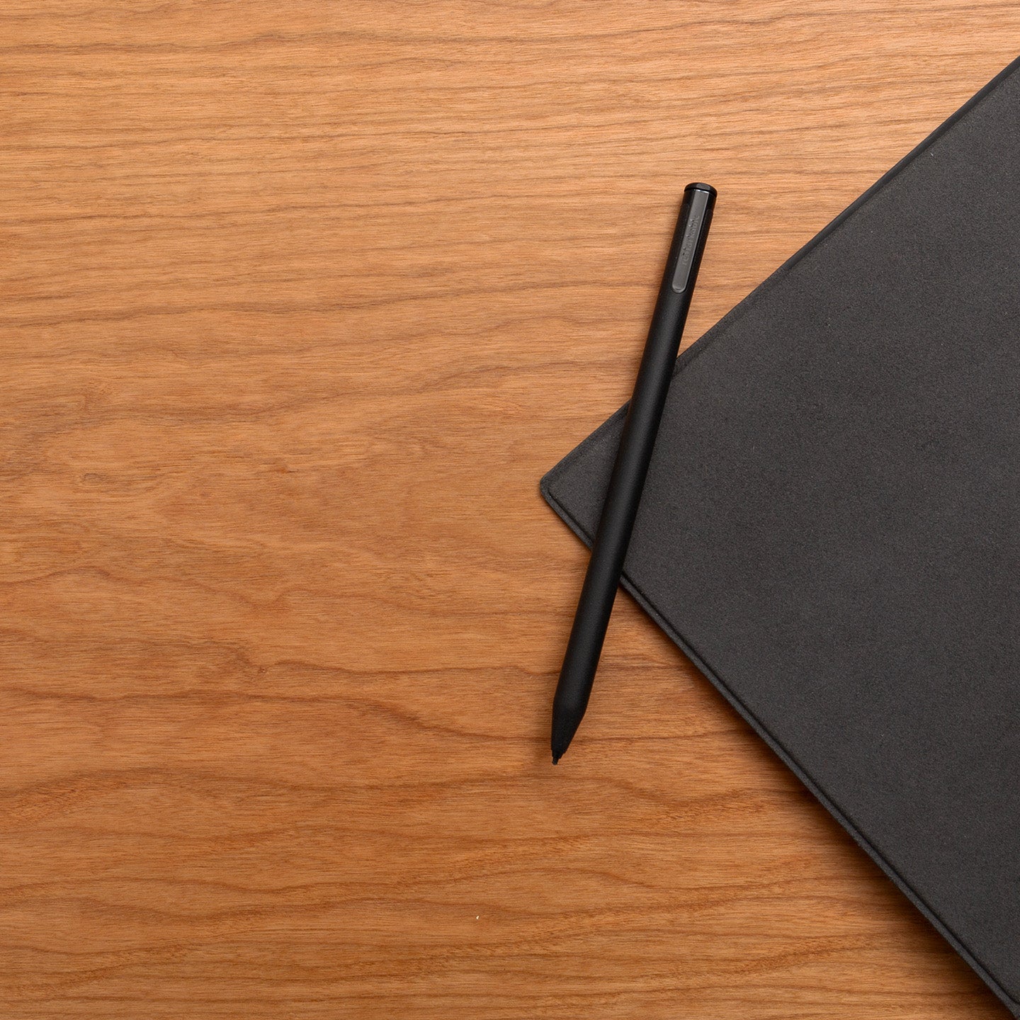 A black pen sits on a closed, dark gray notebook atop a Tischplatte - Furnier Kirsche, both arranged in the upper right corner, leaving most of the vibrant cherry veneer desk surface clear.