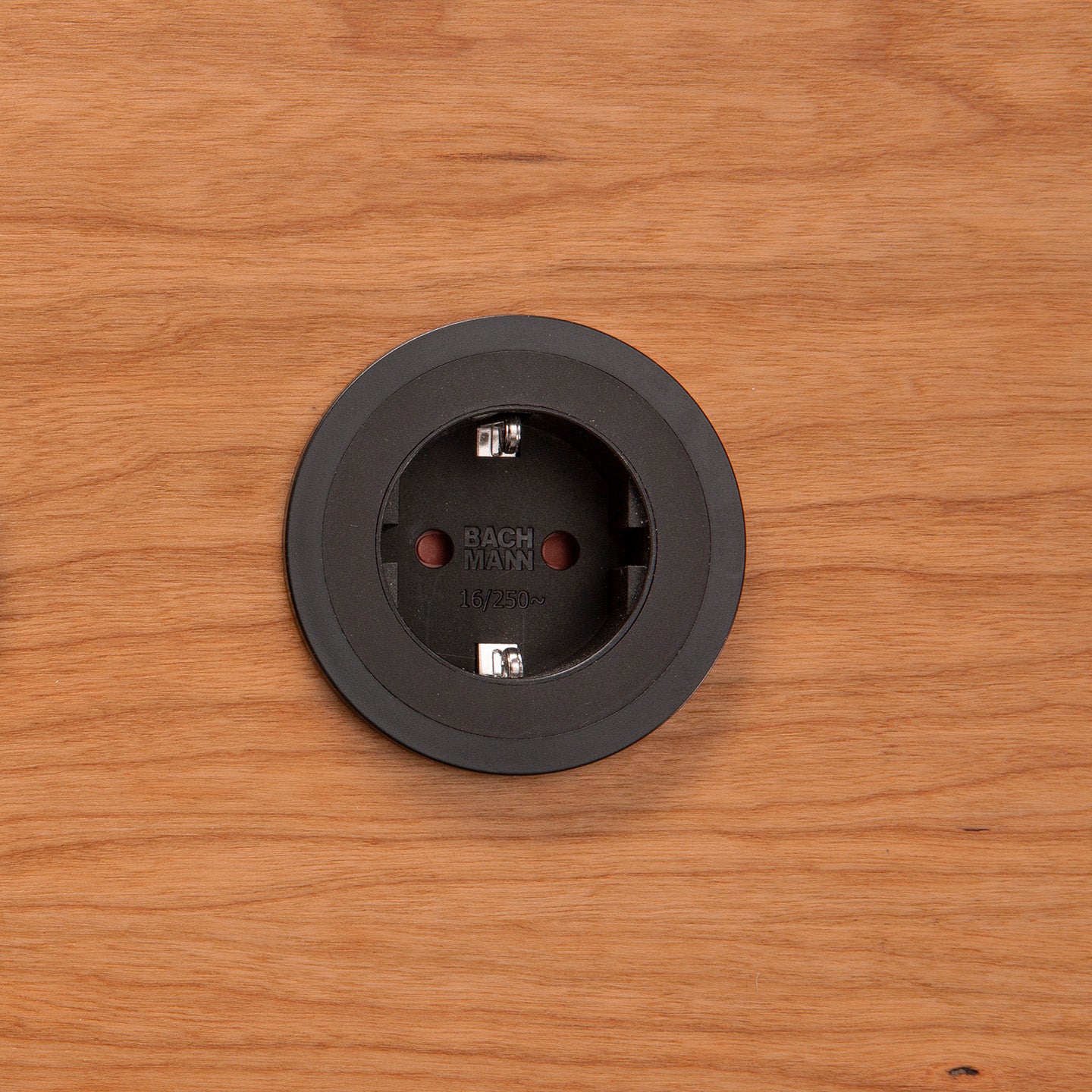 Eine Einbausteckdose Pix in Schwarz ist zentriert in eine Holzoberfläche eingelassen. Die runde Steckdose zeigt zwei Löcher und innen sichtbare Metallkontakte.