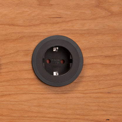 Eine Einbausteckdose Pix in Schwarz ist zentriert in eine Holzoberfläche eingelassen. Die runde Steckdose zeigt zwei Löcher und innen sichtbare Metallkontakte.