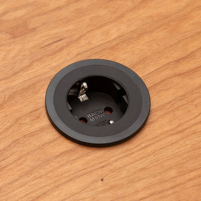 Eine runde, schwarze FOCUS-Steckdose von CUBE Products & Furniture ist von oben gesehen in einen Holztisch eingelassen. Das versenkte Design mit zwei Befestigungsschrauben eignet sich perfekt für kompakte Arbeitsbereiche oder ein modernes Home Office.