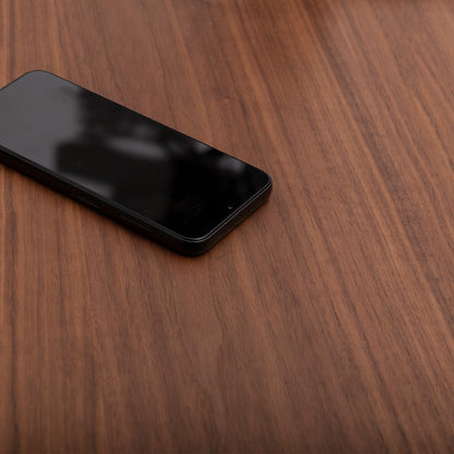 A black smartphone rests face up on the left side of a wooden surface, its dark screen catching the light atop a Tischplatte - Furnier Walnuss, while the elegant walnut veneer table remains empty.