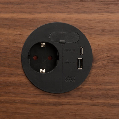 A round black Einbausteckdose & Kombicharger - Loop in Charge with a European socket, USB-C port, and USB-A port is embedded in a wooden surface. The BACHMANN logo appears on the bottom right of the outlet.