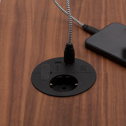 A close-up of a wooden surface shows the Einbausteckdose & Kombicharger - Loop in Charge with a black power outlet and USB port. A phone connects via a black and white braided cable neatly routed through the Kabeldurchführung.