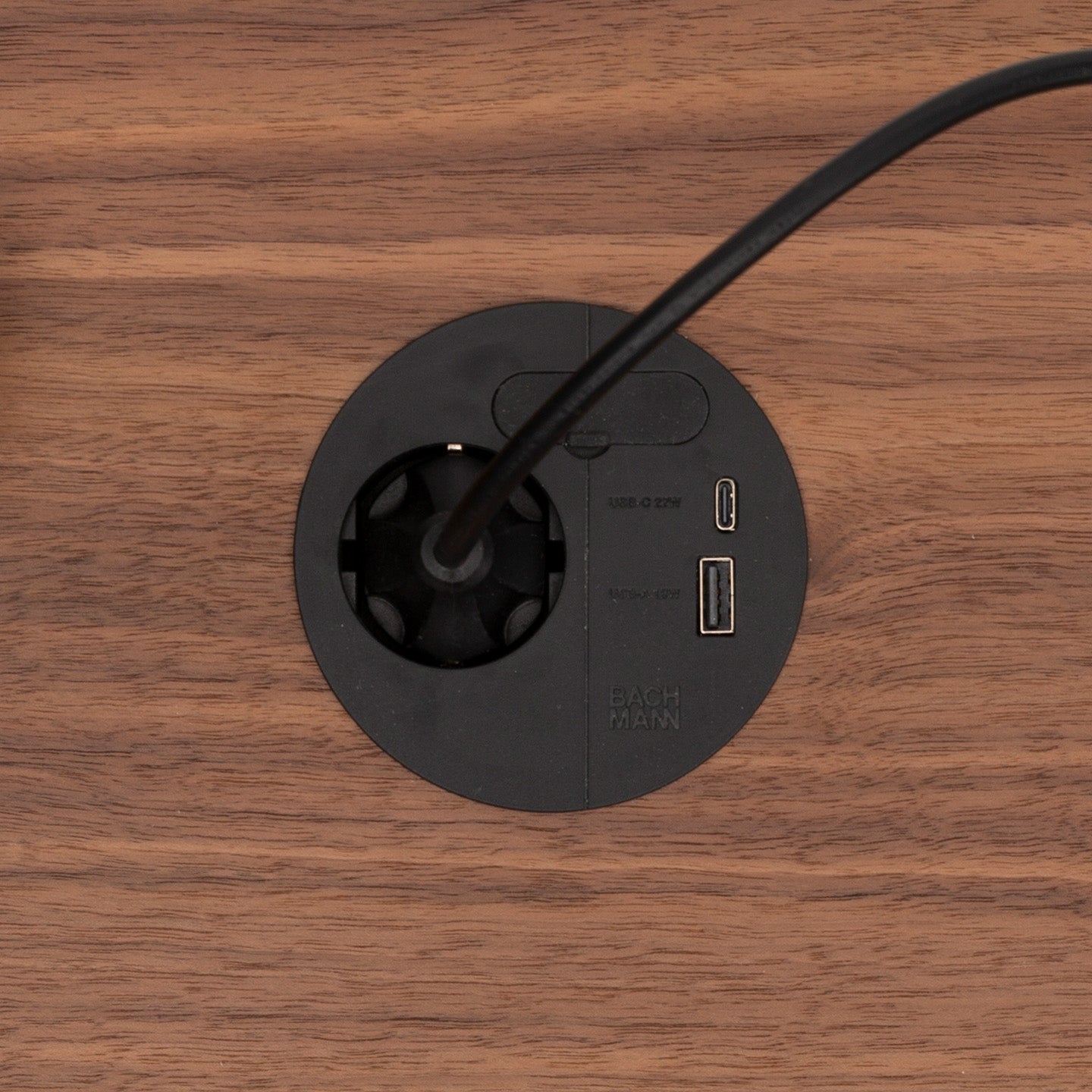 The Einbausteckdose & Kombicharger - Loop in Charge is a black circular power outlet with an attached cable, two USB ports, and a USB-C port, installed in wood and featuring a Kabeldurchführung for organized cables.