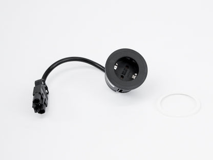 A black round Einbausteckdose Pix with a GST 18 Zuleitung cable and connector rests on a white surface beside a small white circular ring.