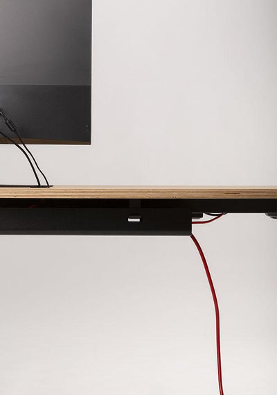 Der MAX-Schreibtisch von CUBE Products & Furniture hat eine Holzoberfläche mit schwarzer Kante, die einen Monitor und sichtbare Kabel (auch rote) trägt und ein XL-Setup und einen organisierten Arbeitsbereich vor einem schlichten Hintergrund hervorhebt.