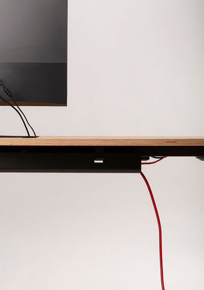 Minimalist side view of a wooden desk with a black monitor partially visible. Red and black cables, organized with the Kabelwanne - 1 Cable PRO, extend from the desk against a light gray background.