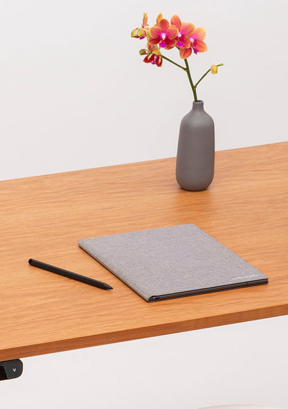 A gray notebook and a black pen rest on a wooden desk, with a small gray vase holding an orange and pink orchid in the background.