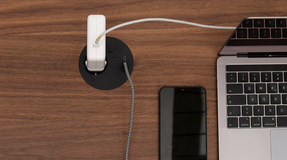 A laptop, smartphone, and charging adapter are placed on a wooden desk. The laptop is partially open, and two cables are plugged into a built-in desk power outlet.